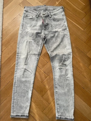 Dsquared2 skinny jeans i grått - Säljer ett par Dsquared2 skinny jeans i grå tvättad färg med slitna detaljer och färgstänk. Modellen är 71630 och har klassisk femficksdesign, knappgylf och Dsquared2-röd etikett. Jeansen är tillverkade i Italien och har en cool, modern look.