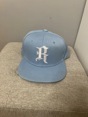 Ljusblå snapback keps med broderat R - Snygg ljusblå snapback keps med platt skärm och ett stort vitt broderat R framtill. 