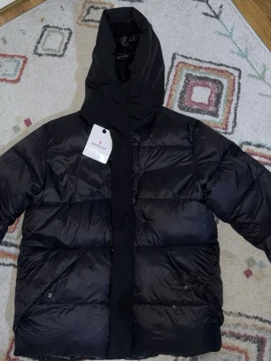 Svart Moncler dunjacka med huva - Snygg svart dunjacka från Moncler med stor huva och quiltad design. Jackan har dragkedja och knappar framtill samt två snedställda fickor med knappstängning. Perfekt för kalla dagar och riktigt trendig look.