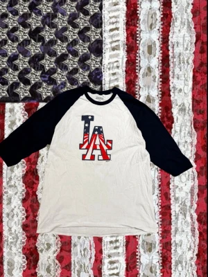 LA baseballtröja med marinblå ärmar - Snygg baseballinspirerad t-shirt från Stitches med vita paneler och marinblå trekvartsärmar. Stor LA-logga framtill i amerikanskt flaggmönster. Perfekt för dig som gillar sportig stil och vill sticka ut.