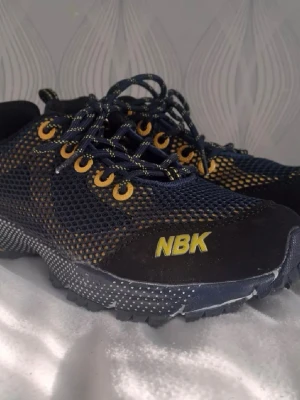 Svarta och gula NBK sneakers - Snygga sneakers från NBK i svart mesh med gula detaljer och snörning. Skorna har en sportig design med robust sula och NBK-logga på sidan. Perfekta för dig som gillar en cool och modern stil.