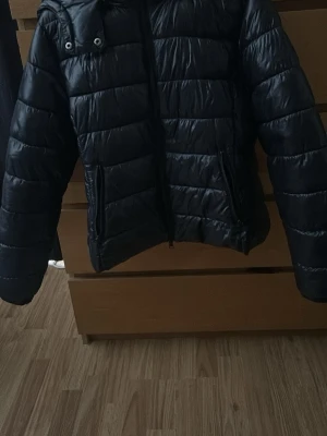 Svart quiltad pufferjacka med huva - Snygg svart pufferjacka med quiltad design och dragkedja framtill. Jackan har huva och långa ärmar, perfekt för kyliga dagar. Tillverkad i polyester för en lätt och bekväm känsla.