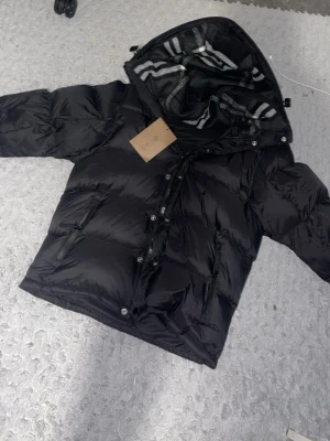 Burberry dunjacka med avtagbara ärmar – 2-i-1 (jacka + väst) - Snygg och stilren Burberry dunjacka i svart, med klassiskt Burberry-rutigt foder i huvan. Jackan har avtagbara ärmar, vilket gör att den även kan användas som dunväst – perfekt för både kalla och mildare dagar.