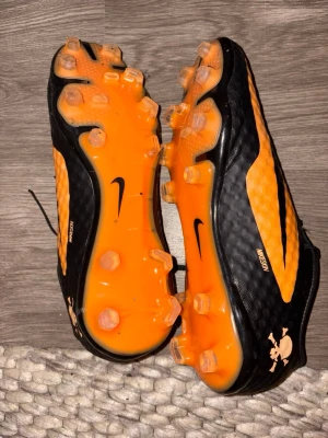Nike orangea och svarta fotbollsskor - Säljer ett par Nike fotbollsskor i svart och orange med snörning och dobbar för gräsplan. Skorna har en sportig design med Nike-logga på sidan och undersidan samt texten Hypervenom på  ovansidan. Tillverkade i syntetmaterial för bra grepp och kontroll.