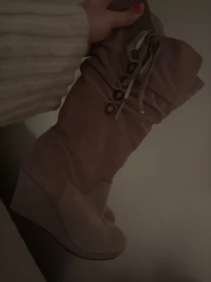 beige mockastövlar med kilklack - säljer ett par snygga beige stövlar i mocka med kilklack och snörning upptill. stövlarna har rund tå och dekorativa knappar längs skaftet. 