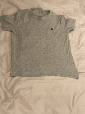Grå t-shirt med U.S. Polo Assn. - Snygg grå t-shirt från U.S. Polo Assn. med klassisk rund hals och korta ärmar. På bröstet finns en liten broderad logga i svart. Materialet är mjukt och bekvämt, perfekt för en avslappnad stil.