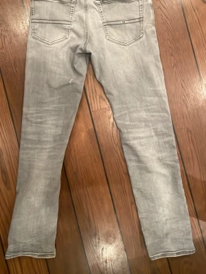 Ljusgrå raka jeans från Tommy Hilfiger - Snygga ljusgrå jeans från Tommy Hilfiger med raka ben och klassisk femficksdesign. Jeansen har diskreta slitningar och logodetaljer på bakfickan. Perfekta för en avslappnad och trendig look. Tillverkade i mjuk bomullsdenim.