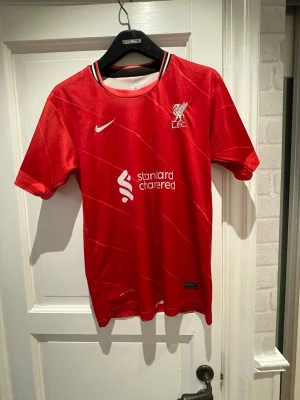 Röd Liverpool Nike fotbollströja  - Liverpool FC matchtröja från Nike med Dri-FIT material, röd med vita detaljer och klubbmärke på bröstet. Nummer 11 och M. Salah på ryggen. Klassisk krage med vita ränder. Perfekt för dig som älskar fotboll och vill bära ditt favoritlag.