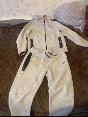 Grå Nike huvtröja med dragkedja - Säljer en grå huvtröja från Nike med hel dragkedja framtill och svarta detaljer vid fickorna. Tröjan har en sportig look med huva och är tillverkad i mjukt material, perfekt för chill eller träning. Klassisk Nike-logga på bröstet. Den har ett litet hål på benet