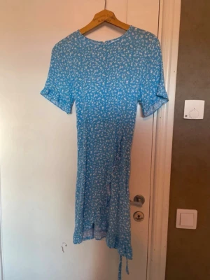 Blommig blå klänning från Vila - Supersöt blå klänning från Vila med vitt blommönster. Klänningen har korta ärmar, rund hals och en omlott-detalj med knyt i sidan. Perfekt för sommarens alla tillfällen.