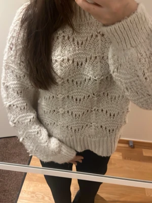 stickad tröja med detaljer - Mysig chunky stickad tröja i beige med hålmönster och breda ribbade muddar vid ärmslut och nederkant. Tröjan har en avslappnad passform och hög hals, perfekt för kalla dagar. Materialet ser ut att vara en mjuk ull- eller ullmix.
