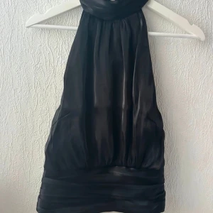 Svart halterneck topp i satin - Snygg svart halterneck topp i glansigt satinmaterial. Toppen är ärmlös och har en hög hals med bred kant nedtill som ger en elegant siluett. Perfekt för dig som vill ha en stilren och trendig look.
