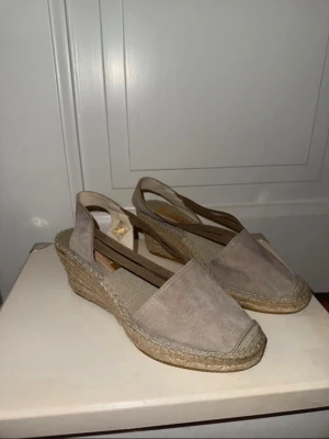 Beige kilklackade sandaler i mocka - Snygga beige sandaler med kilklack och öppen häl. Ovandelen är i mjuk mocka och sulan har en klassisk flätad espadrille-look. Elastiska band över foten för skön passform. Perfekta till sommaren och ger en avslappnad men chic vibe. Klackhöjd 7,5