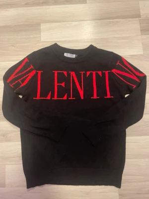 Svart stickad tröja från Valentino - Svart stickad tröja från Valentino med stora röda bokstäver över bröstet och ärmarna. Tröjan har rund halsringning och ribbade muddar vid ärmslut och nederkant. Snygg och statement med tydlig logga framtill.