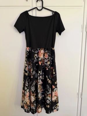 Svart blommig midiklänning - Supersöt svart midiklänning med ribbad överdel och korta ärmar. Kjolen har ett färgstarkt blommönster i rosa, beige och vitt som ger en romantisk vibe. Perfekt för sommarens alla tillfällen! Materialet känns mjukt och följsamt.