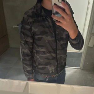 Camouflage vindjacka från SOHO NYC - Snygg vindjacka i mörkgrått och svart camomönster från SOHO NYC. Jackan har dragkedja framtill, hög krage och elastiska muddar vid ärmslut och midja. 