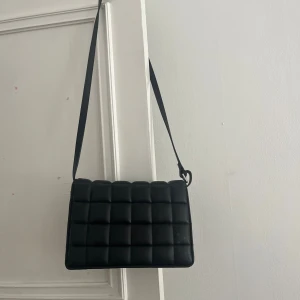 Svart quiltad axelväska i skinnimitation - Stilren svart axelväska med quiltad design och lång justerbar rem. Väskan har en rektangulär form och är tillverkad i skinnimitation. Perfekt storlek för det viktigaste och med innerfack för extra förvaring.