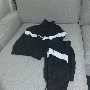 Svarta träningskläder - Svarta täckbyxor med bred vit diagonal rand över benen. Byxorna har resår i midjan och vid bensluten samt snörning i midjan för extra passform. Materialet är syntet och ger en sportig vibe, perfekt för utomhusaktiviteter.