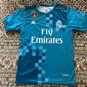 Real Madrid Ronaldo 7 bortatröja Adidas - Säljer en blå Real Madrid bortatröja från Adidas med Ronaldo 7 på ryggen. Tröjan har korta ärmar, CL-märken på ärmarna och Fly Emirates-tryck på bröstet. Tillverkad i lätt och ventilerande Climacool-material. Snygga pixelrutor framtill och broderad klubbmärke.