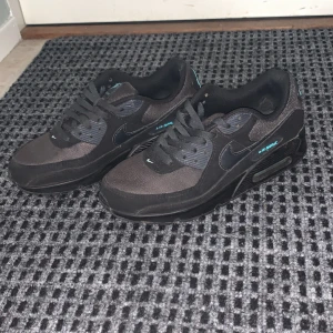 Nike Air Max 90 svart med turkos - Säljer ett par svarta Nike Air Max 90 sneakers med turkosa detaljer på loggan och tungan. Skorna har klassisk siluett, snörning och en blandning av textil och syntetmaterial. Perfekta för dig som gillar streetstyle och vill ha en bekväm sneaker med snygg design. Köpta för 2000:-
