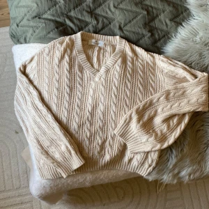 Beige kabelstickad tröja från H&M - Kabelstickad beige tröja från H&M med v-ringning och breda ribbade muddar. Tröjan har ett klassiskt flätmönster och är tillverkad i mjuk bomull. Perfekt för lager-på-lager och chill höst- och vinterstil.