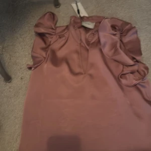 Rosa blus med voluminösa ärmar Neo Noir - Supersnygg rosa blus från Neo Noir i glansigt satinmaterial. Blusen har voluminösa, korta puffärmar och en v-ringad halsringning. Perfekt för dig som vill sticka ut med en trendig och feminin look. Färgen är en mjuk, dammig rosa och materialet känns lyxigt.