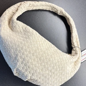 Beige axelväska i mocka med flätad look - Trendig beige axelväska med flätad struktur och mjuk bågform. Väskan är gjord i mocka och har en snygg guldfärgad dragkedja upptill. Perfekt storlek för att bära med sig det viktigaste och ger en cool touch till din outfit.