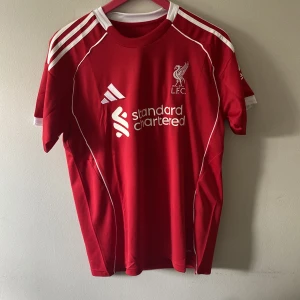 Liverpool FC röd Adidas fotbollströja - Liverpool FC fotbollströja från Adidas i rött med vita detaljer och broderad LFC-logga på bröstet. Tröjan har klassiska Adidas-ränder på axlarna, vit krage och sponsortryck framtill. Tillverkad i lätt och ventilerande material.
