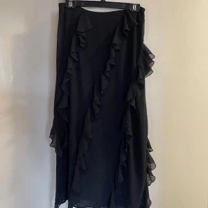 Lång svart volangkjol med slits *OANVÄND* - En superfin långkjol från Lindex i storlek 38 ❣️ Den sitter perfekt i längden på mig som är strax över 170 cm lång. Skulle säga att det är en medelhög/låg midja. Kjolen har en slits och en förslutning med dragkedja vid sidan om höften. Var jättenöjd när jag köpte den men det blev att jag aldrig använde den. 