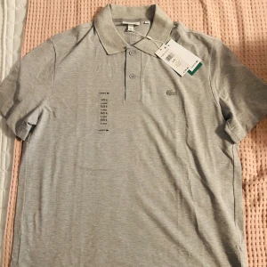 Grå pikétröja från Lacoste XL - Snygg grå pikétröja från Lacoste i storlek XL. Klassisk krage, knappslå med två knappar och den ikoniska krokodilloggan på bröstet. Tillverkad i en mjuk mix av bomull och polyester för skön passform och stilren look.