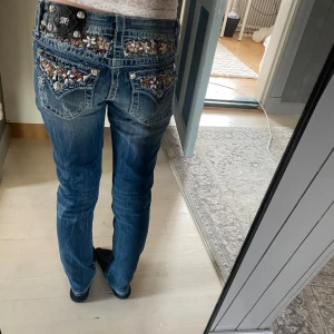 Blå jeans från Miss Me - Snygga blå jeans från Miss Me med broderade detaljer och blommönster på bakfickorna. De har en lågmidjad passform och är dekorerade med knappar och dragkedja. Perfekta för dig som gillar en unik stil.  