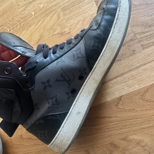 Louis Vuitton svarta high-top sneakers - Säljer ett par svarta high-top sneakers från Louis Vuitton med klassiskt monogrammönster. Skorna har snörning, bred rem med LV-logga vid ankeln och vit platt sula. Tillverkade i skinn med detaljer i textil och gummi. Riktigt snygga och exklusiva sneakers för dig som gillar streetstyle. Äkta!