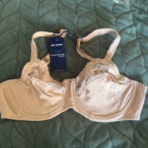 Beige bh från Fantasie - Supersnygg beige bh från Fantasie med vackra spetsdetaljer och broderade partier upptill. Modellen har justerbara axelband och en liten hängande berlock framtill. Materialet är mjukt och skönt med diskret mönster i tyget.