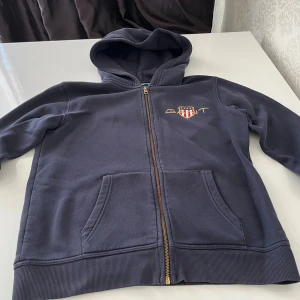 Mörkblå hoodie från GANT med dragkedja - Snygg mörkblå hoodie från GANT med huva och hel dragkedja framtill. Tröjan har två fickor och ett broderat GANT-emblem på bröstet. Tillverkad i mjuk bomull, perfekt för chill dagar. Klassisk passform och långa ärmar.