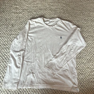 Vit långärmad tröja från Polo Ralph Lauren - Vit långärmad tröja från Polo Ralph Lauren med klassisk rund hals och broderad marinblå logga på bröstet. Tillverkad i mjuk bomull, perfekt för en clean och enkel stil. Passar dig som gillar basic men ändå vill ha något från ett ikoniskt märke.