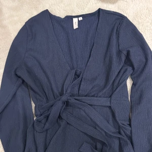 Mörkblå blus  från Nelly - Mörkblå blus från Nelly med lång ärm och knytband i midjan. Blusen har v-ringning och är tillverkad i ett lätt, strukturerat tyg som ger en snygg och modern look. Perfekt för dig som gillar stilrena och enkla plagg med en twist.