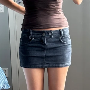 Svart jeansminikjol med låg midja - Säljer en svart jeansminikjol med låg midja och klassiska femficksdetaljer. Kjolen har bälteshällor, knapp- och dragkedjestängning framtill och en tight passform. 