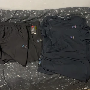 Svart träningsshorts från Under Armour 2 sets  - Säljer ett par svarta träningsshorts från Under Armour i storlek M. Shortsen har elastisk midja med snörning och en diskret logga i regnbågsfärger på ena benet. Tillverkade i ett lätt och snabbtorkande syntetmaterial som passar perfekt för träning.
