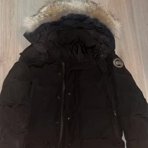 Svart dunjacka Canada Goose med pälskrage - Säljer en svart dunjacka från Canada Goose med fluffig pälskant på huvan och klassisk patch på ärmen. Jackan har dragkedja, knappar och flera fickor. Perfekt för kalla dagar 