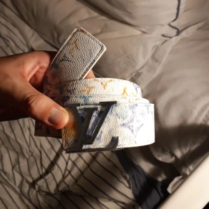 Louis Vuitton vitt bälte med färgstänk - Säljer ett Louis Vuitton bälte i vitt skinn med coola färgstänk i blått, orange och gult. Bältet har en ikonisk LV-spänne i silver och präglad Louis Vuitton-logga på baksidan av spännet. Perfekt för dig som vill sticka ut med en unik accessoar.