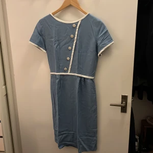 Ljusblå kortklänning med vita detaljer - Supersöt ljusblå klänning med vita kanter och dekorativa knappar framtill. Klänningen har korta ärmar, rund halsringning och markerad midja. Stängs med dragkedja bak. Perfekt för dig som gillar retrovibbar och stilrena detaljer.💖