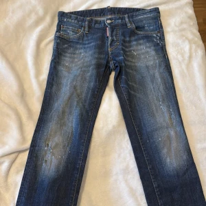 Blå jeans från Dsquared2, stl 44 - Säljer ett par blå jeans från Dsquared2 i storlek 44. Jeansen har en snygg tvätt med slitningar och färgstänk, klassisk femficksmodell och tydliga kontrastsömmar. Märkeslapp på bakfickan och röd Dsquared2-tag vid gylfen. Perfekta för en cool streetstil.