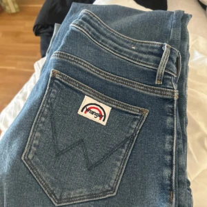 Wrangler Flare jeans blå W27 L30 - Säljer ett par blåa Wrangler jeans med flare-ben och klassiska fickor både fram och bak. Jeansen har medel hög midja och är tillverkade i mjukt denimtyg. Snygg detalj med Wrangler-loggan på bakfickan. Perfekta för dig som gillar retrovibbar och bekväm passform.