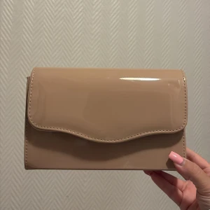 Beige kuvertväska med kedja - Snygg beige kuvertväska i glansigt material med stilren design och vågig kant på locket. Väskan har en tunn guldfärgad kedja som axelrem, perfekt för att lyfta din outfit. Enkel att matcha och rymmer det viktigaste du behöver ha med dig.