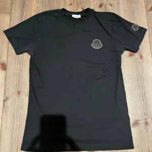 Svart Moncler t-shirt med logga - Svart t-shirt från Moncler med klassisk rund hals och korta ärmar. Stora svarta Moncler-loggor med orange kant sitter på bröstet och ena ärmen. T-shirten är i mjuk bomull och har en clean, stilren look som passar perfekt till streetwear.