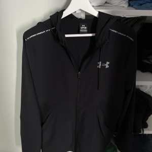 Svart jacka från Under Armour - Svart jacka från Under Armour med dragkedja och huva. Snyggt logotryck på bröstet och textdetaljer på axlarna. Perfekt för träning eller chill. Tillverkad i mjukt syntetmaterial som andas. Långärmad och med praktiska fickor framtill.