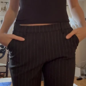 Svarta kostymbyxor med smala ränder - Snygga svarta lågmidjade kostymbyxor med tunna vita ränder och klassisk passform. Byxorna har hällor för bälte, raka ben och normal midja. Perfekta för en stilren look och passar till både sneakers och boots.