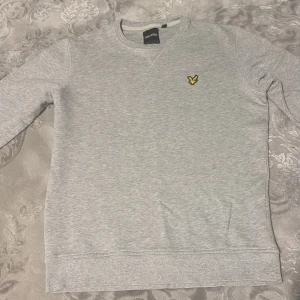 Grå sweatshirt från Lyle & Scott - Klassisk ljusgrå sweatshirt från Lyle & Scott med rund halsringning och broderad gul logga på bröstet. Tröjan är i mjuk bomull och har ribbade muddar vid ärmslut och nederkant. Perfekt för en chill och stilren look.