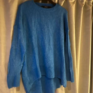 Blå stickad tröja från Vero Moda - Mysig blå stickad tröja från Vero Moda i storlek S. Tröjan har en loose passform, rund halsringning och långa ärmar med breda muddar. Perfekt för lager-på-lager och chill dagar. Snyggt längre baktill för en avslappnad look.
