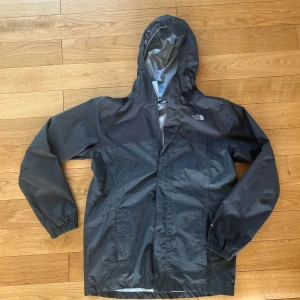 Svart/grå vind/regnjacka från The North Face - Snygg vindjacka från The North Face i svart och mörkgrått. Jackan har huva, dragkedja framtill och två sidofickor. Tillverkad i lätt och slitstark polyester, perfekt för blåsiga dagar. Logga på bröstet och skuldran med elastiska muddar vid ärmsluten. Storlek: Boys xl, motsvarar en Small herr